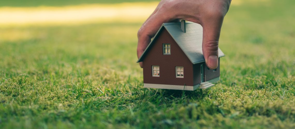 Concept of selling a house. A hand is holding a model house above green meadow.