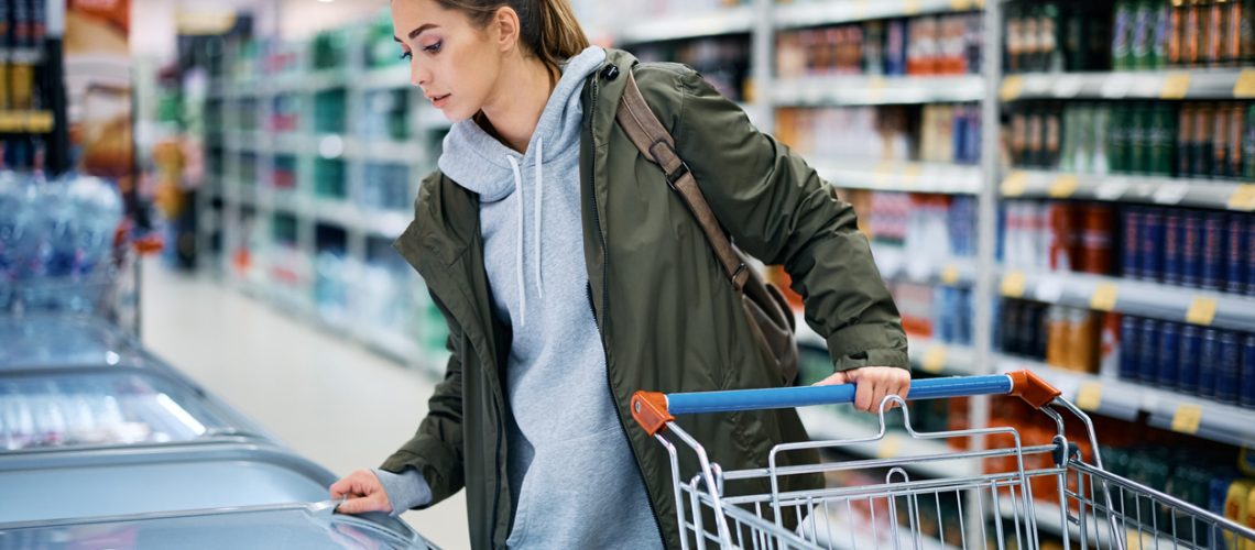 Young woman looking at frozen products in freezer while shopping in grocery store.